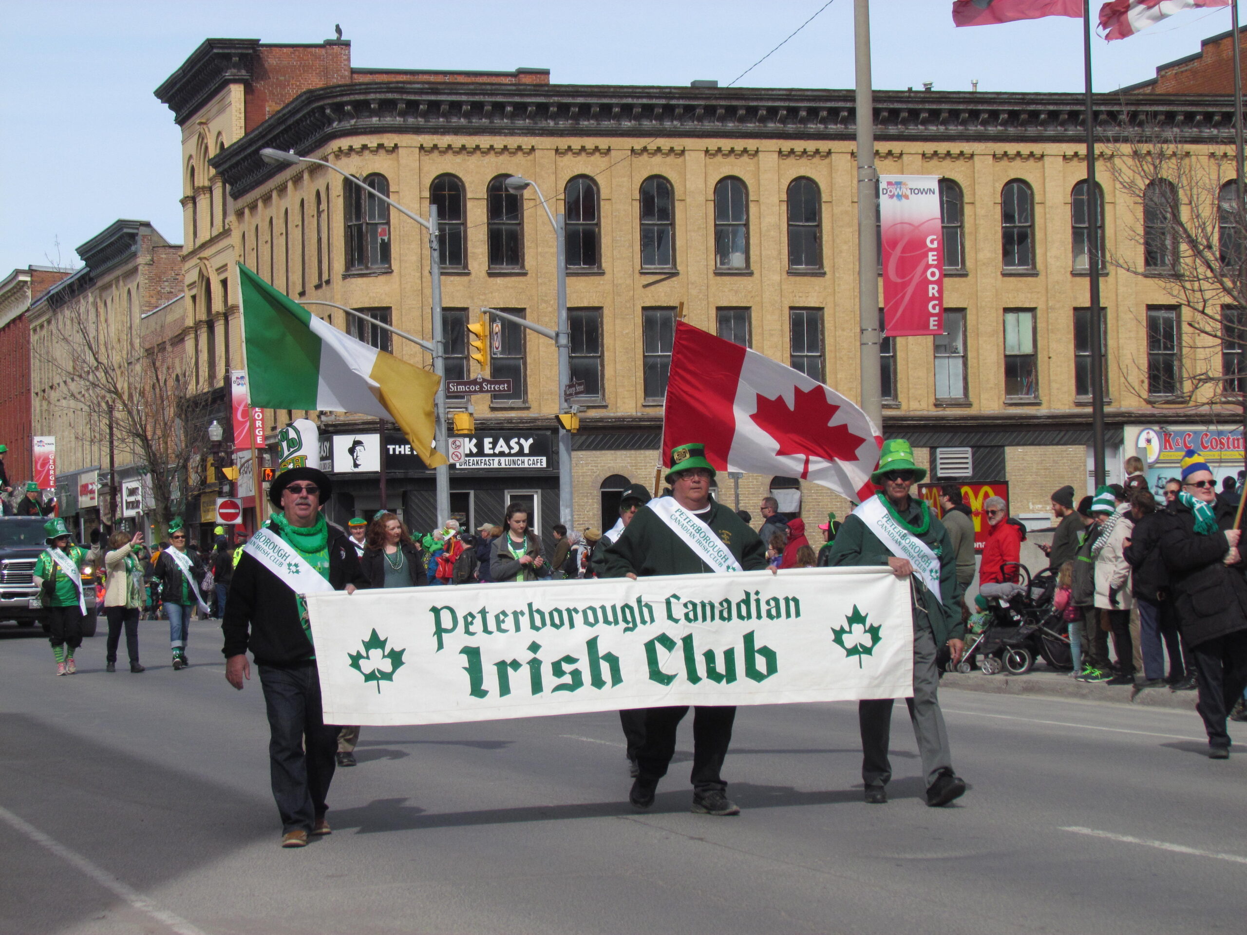 A group of people are marching in a parade, carrying a banner that reads "Peterborough Canadian Irish Club." They are holding Irish and Canadian flags, and many are wearing green hats. Brick buildings and spectators are visible in the background.