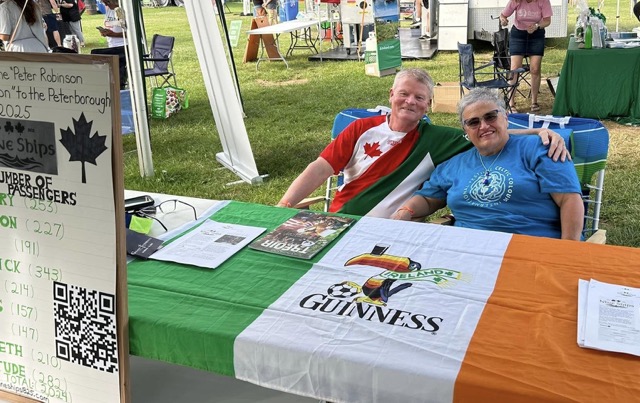 Jay and Rose sitting behind the booth of of Nine Ships Inc. during the Niagara Irish Festival. The table is adorned with the Irish flag and information about the bicentennial.