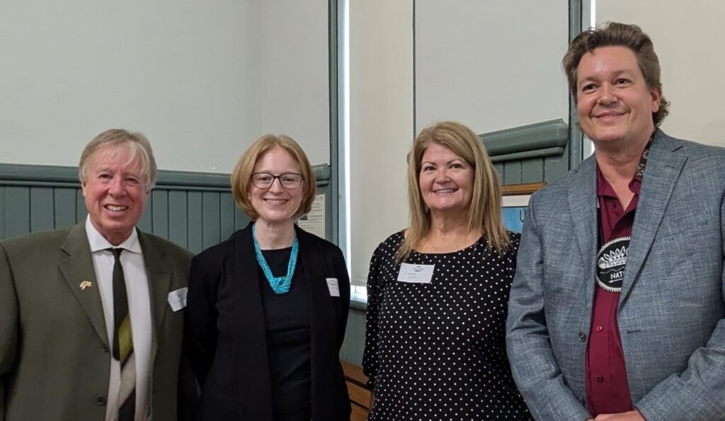 Brendan Moher, Janice McGann, Chief Cowie, Chief Simpson stand next together with smiles, the background is beige and green with a window. 