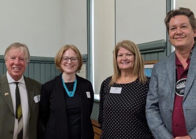 Brendan Moher, Janice McGann, Chief Cowie, Chief Simpson stand next together with smiles, the background is beige and green with a window.