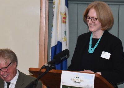 Brendan Moher sits to the left and laughs as Janice McGann (right) stands at the podium and makes a joke in her speech. The background is wood panelling with the county flag in view.