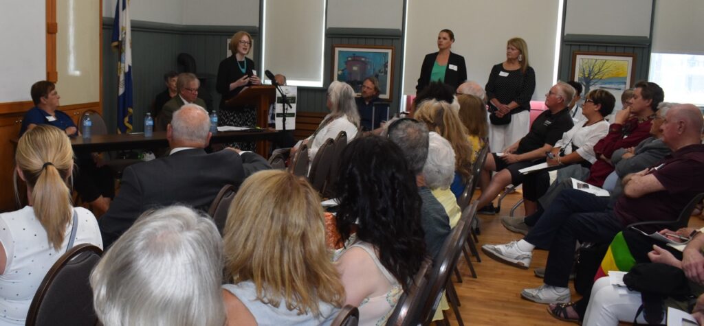 Media photo overlooking the crowd in a room, in the background the Consul General stands at a podium to speak. 