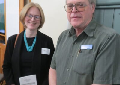 Janice McGann stands next to Don Willcock posing for a picture, the background is wood panelling and a beige wall.