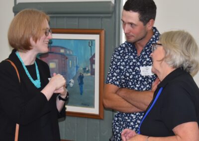 From left to right: Janice McGann with Patrick Leahy and Mary Claire Moher into conversation, the background features a painting of a train car hanging on a wood panel wall.