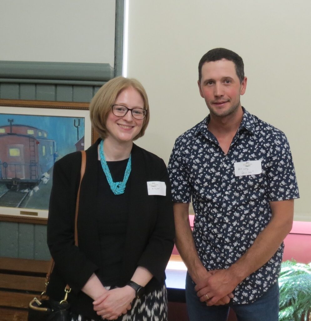 Consul General Janice McGann stands next to Patrick Leahy with smiles. In the background on the left a painting of a train car, a covered window and a small fern in the right corner. 