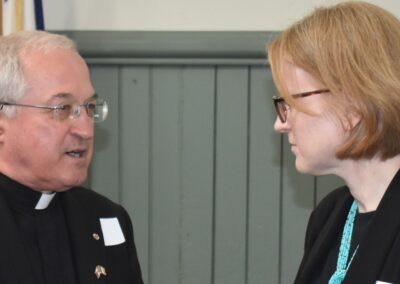 Father Tom on the left chats with Janice McGann on the right. In the background is basic wall of greenish-grey wood panelling.