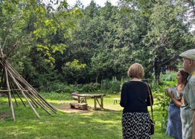 Janice McGann, Ruth O'Connell and Elwood Jones stand facing the Aabnaabin Camp display of a teepee base structure and camp featured outdoors at the Lang Pioneer Village Museum. The background is lush green forest.