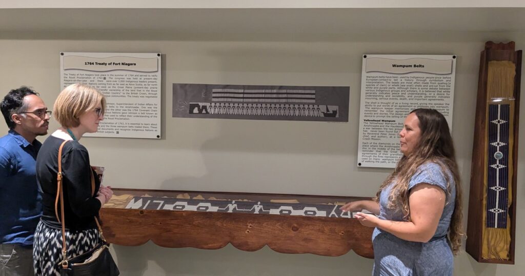 Ruth O'Connell on the right of the Aabnaabin Exhibit explaining, while Consul General Janice McGann and Daniel Capistrano on the right observe their explanation. 