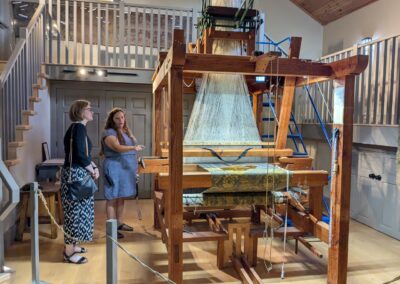 Ruth O'Connell showing off the Jacquard Loom exhibit (a very large wooden loom) in the centre of one of the museum rooms to Janice McGann at Lang Pioneer Village.