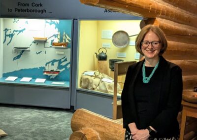 Janice McGann stands in front of a large Peter Robinson exhibit display featuring log cabin structure and a journey map with replica boats in the background.